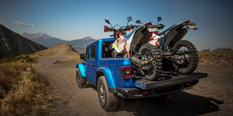 A blue 2025 Jeep Gladiator pickup truck parked on a dirt road, carrying two dirt bikes in the back.