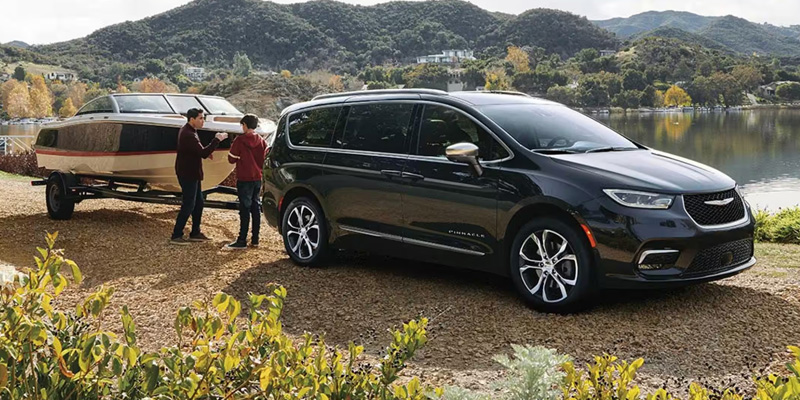 A black Chrysler-Pacifica is parked by a lake, towing a boat. Two people are beside the boat on gravel, set against lush green hills and a cloudy sky.